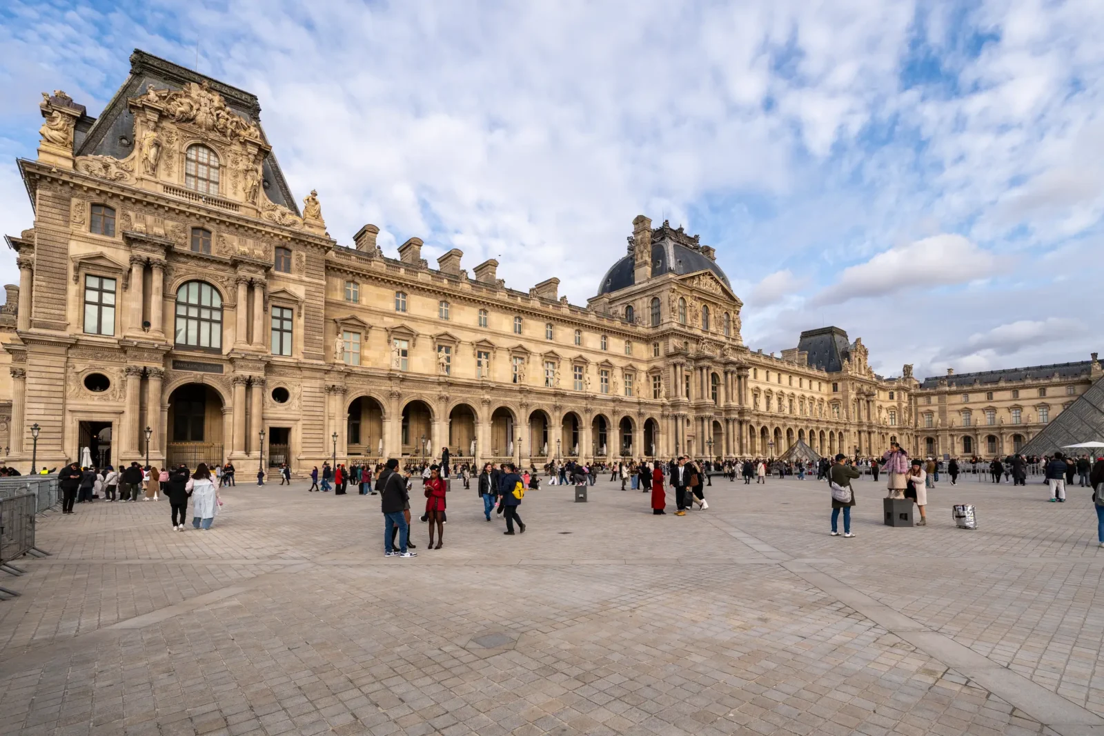 Le majestueux Palais du Louvre et sa cour pavée animée sous un ciel nuageux.