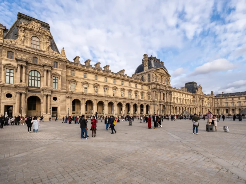 Le majestueux Palais du Louvre et sa cour pavée animée sous un ciel nuageux.