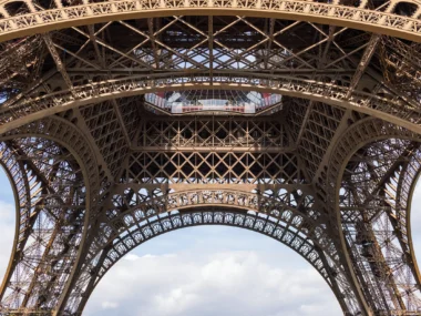 Vue d'en bas de la Tour Eiffel, révélant sa structure métallique complexe sous un ciel nuageux.