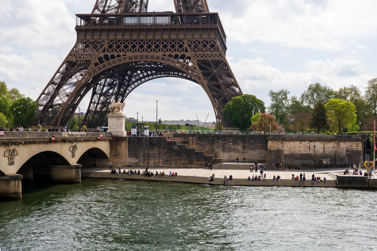 La Tour Eiffel surplombe la Seine où des gens se reposent sur les marches ensoleillées.