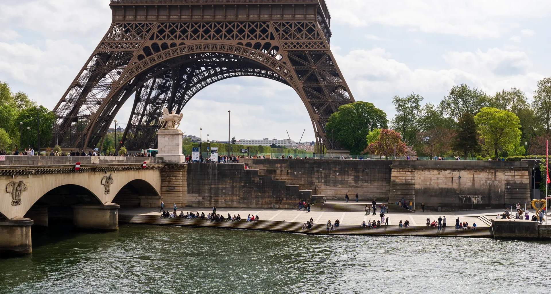 La Tour Eiffel surplombe la Seine où des gens se reposent sur les marches ensoleillées.