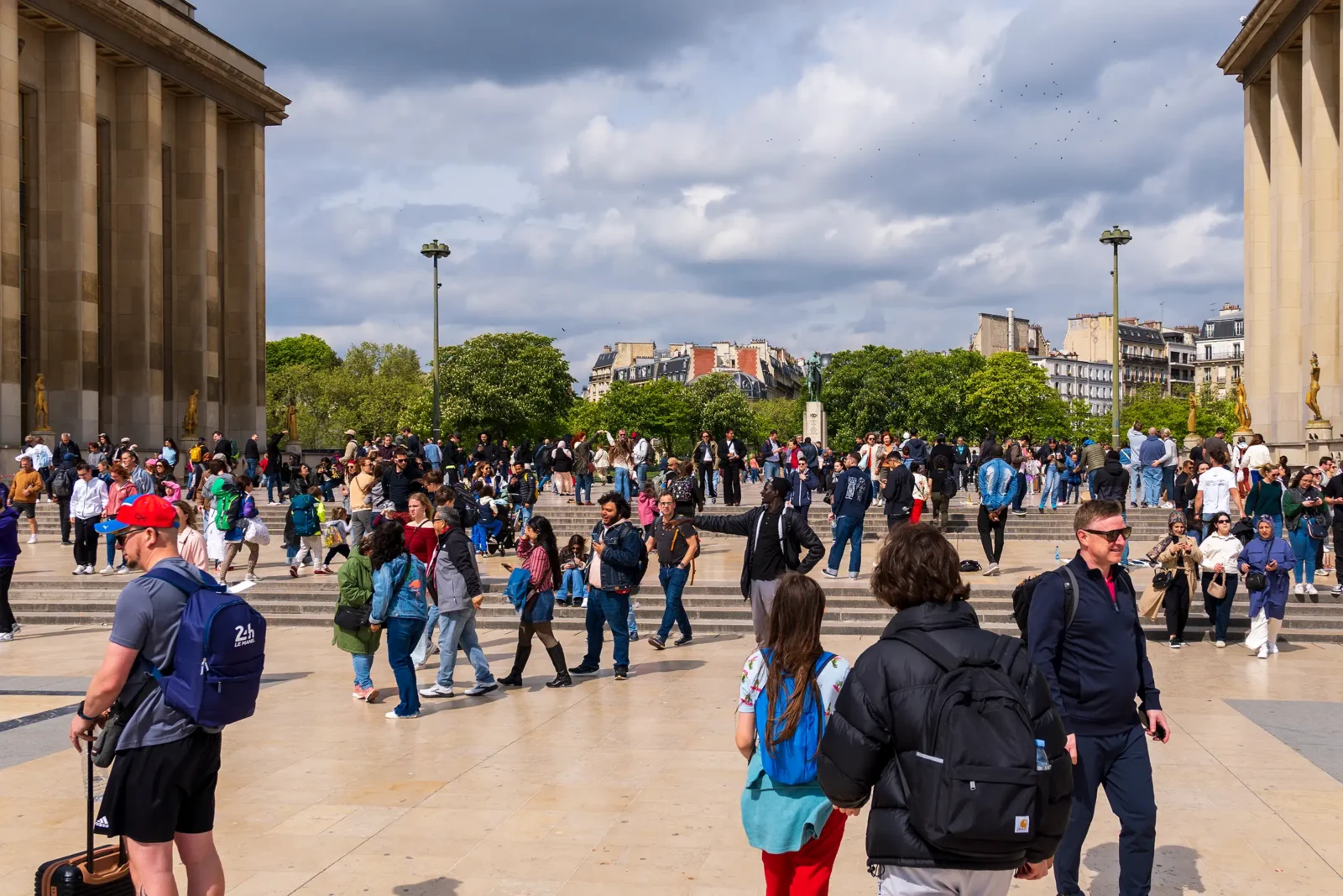 Foule de personnes sur une place ensoleillée devant un bâtiment historique et des arbres.