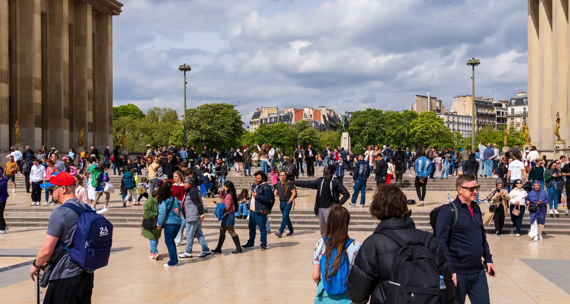 Foule de personnes sur une place ensoleillée devant un bâtiment historique et des arbres.