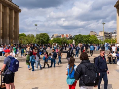 Foule de personnes sur une place ensoleillée devant un bâtiment historique et des arbres.