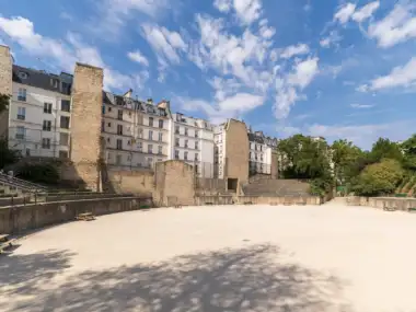 Vue ensoleillée des anciennes Arènes de Lutèce bordées par des immeubles parisiens.