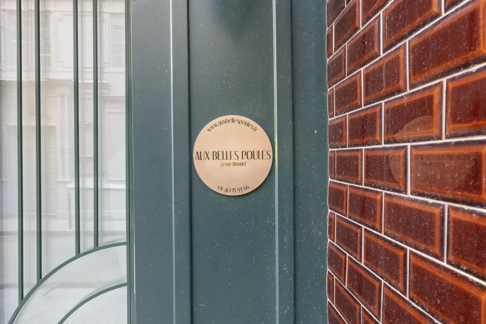 Une plaque ronde dorée Aux Belles Poules est fixée sur une porte verte parisienne.