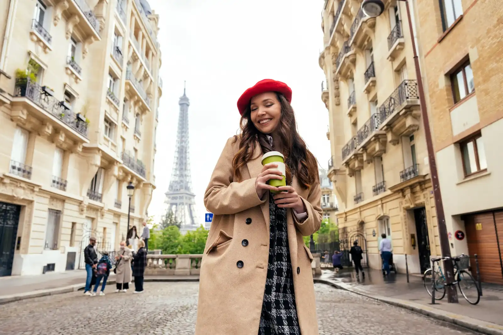 Une jeune femme souriante en manteau beige et béret rouge tient un gobelet de café à Paris.