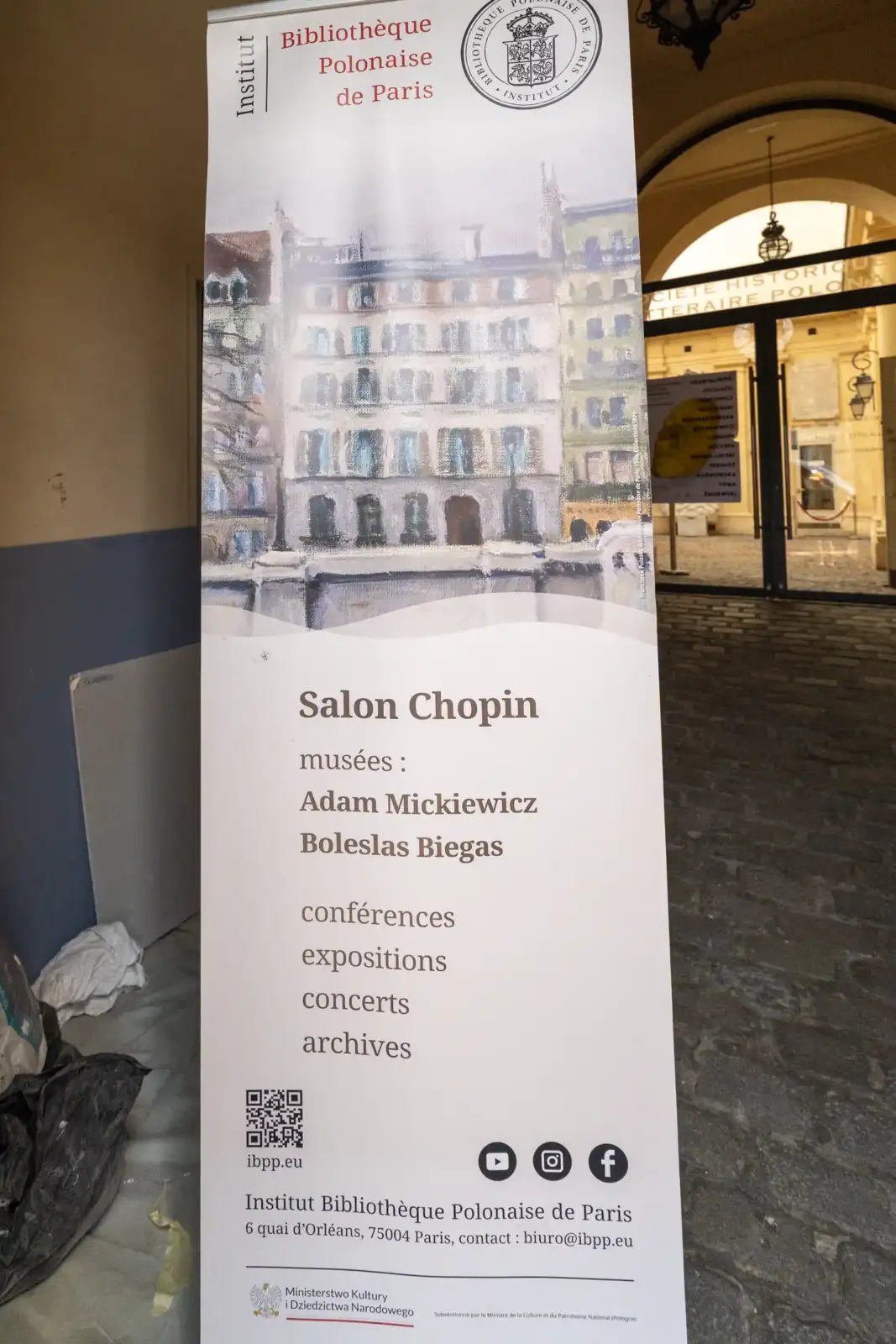Un panneau blanc vertical de la Bibliothèque Polonaise de Paris est dressé dans une cour.