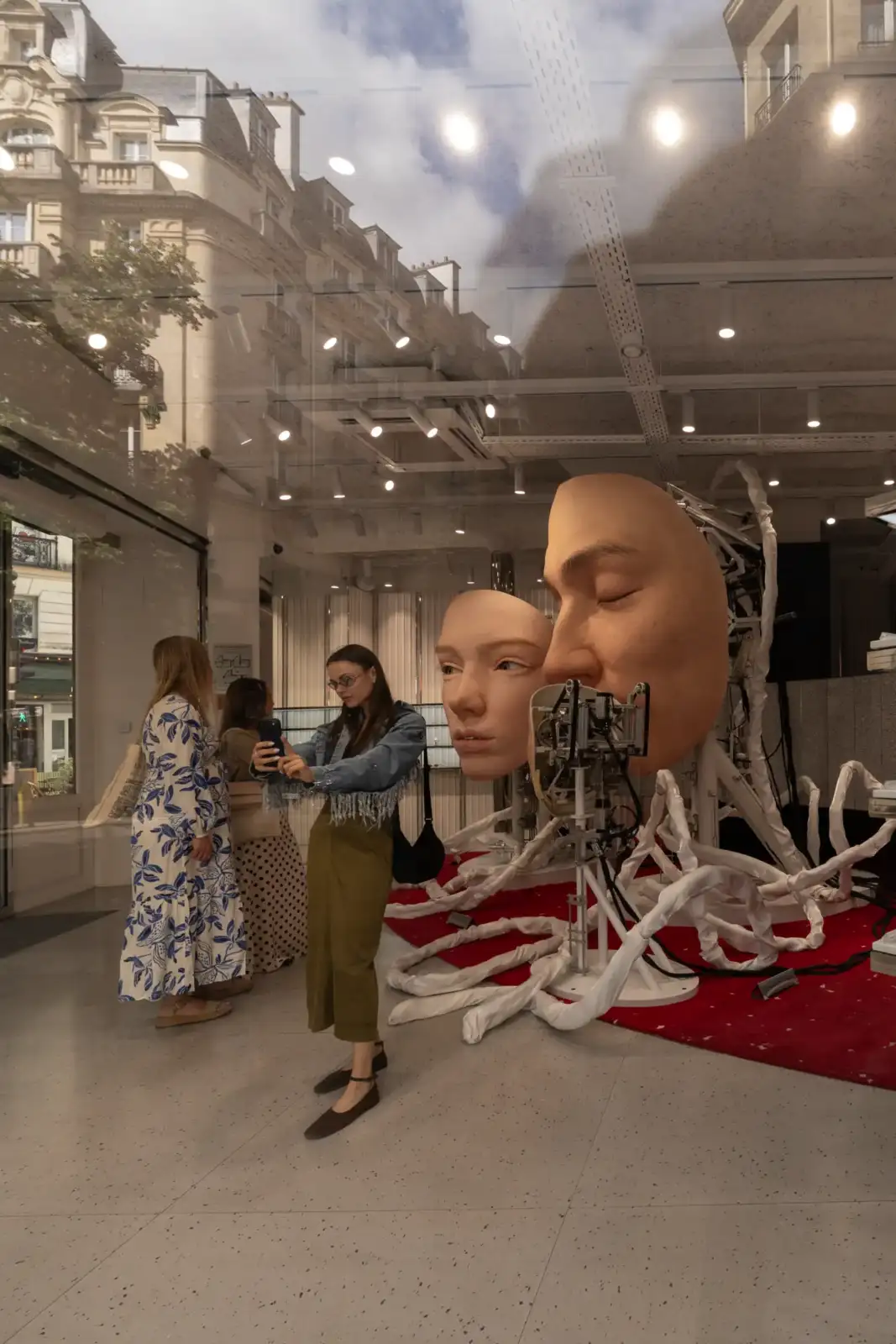 Une femme prend en photo deux immenses têtes robotisées exposées dans une vitrine parisienne.
