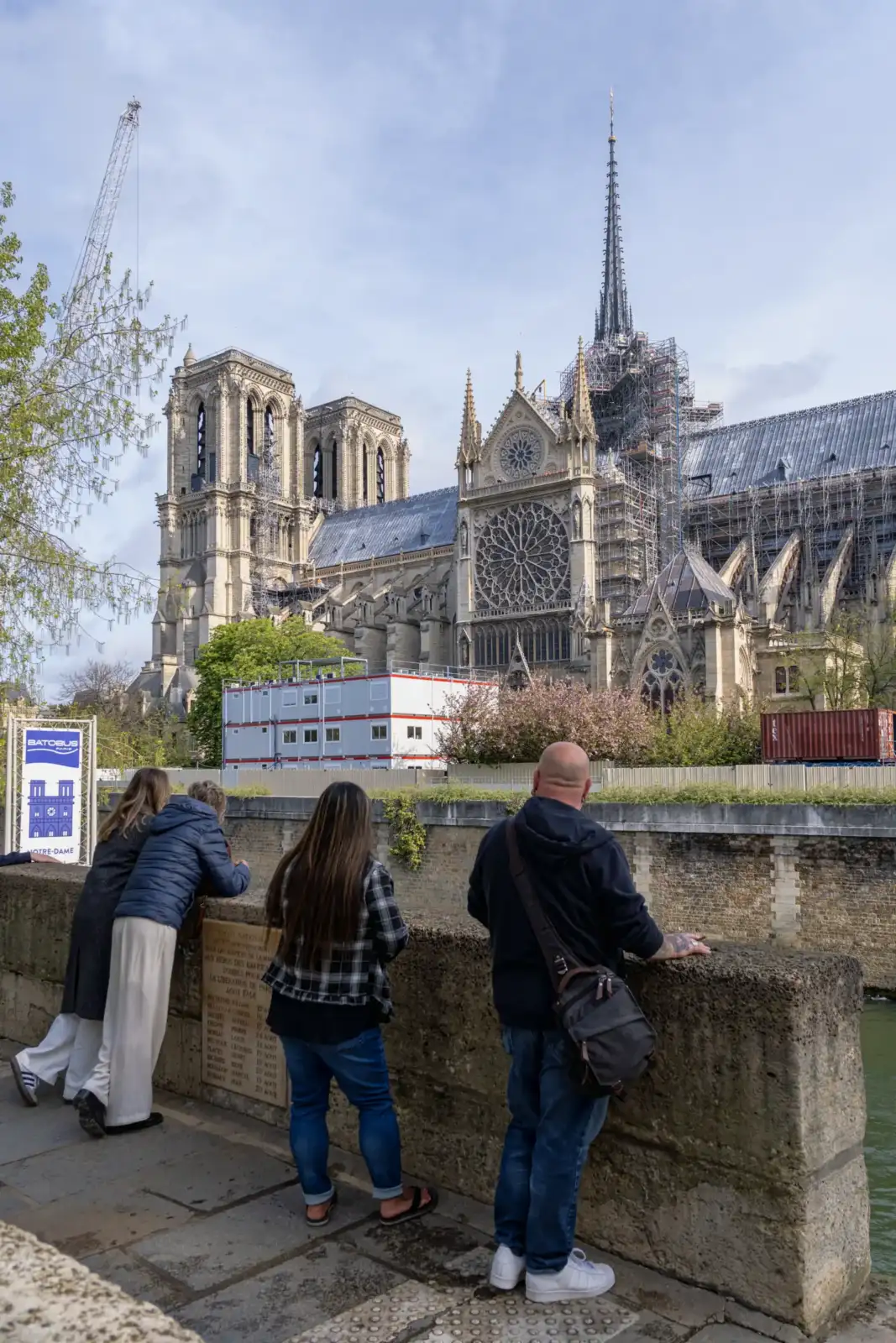 Des touristes observent la cathédrale Notre-Dame de Paris en pleine restauration depuis un quai.
