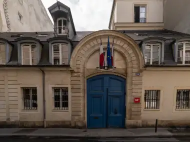La façade classique de l’immeuble beige arbore une grande porte bleue et des drapeaux.