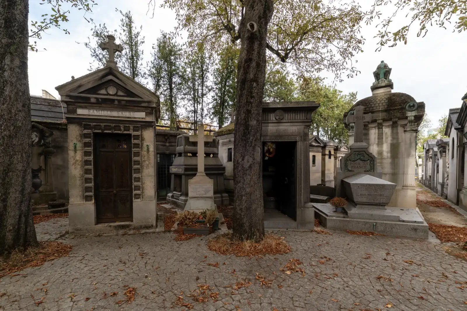 Des caveaux funéraires en pierre et des arbres ombragent une allée pavée du cimetière.