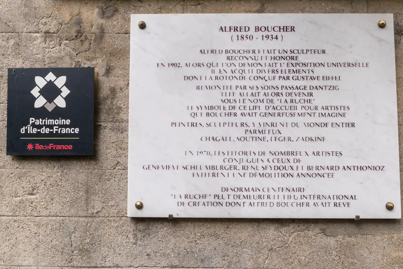 Une plaque commémorative en marbre blanc fixée sur un mur de pierre brute.