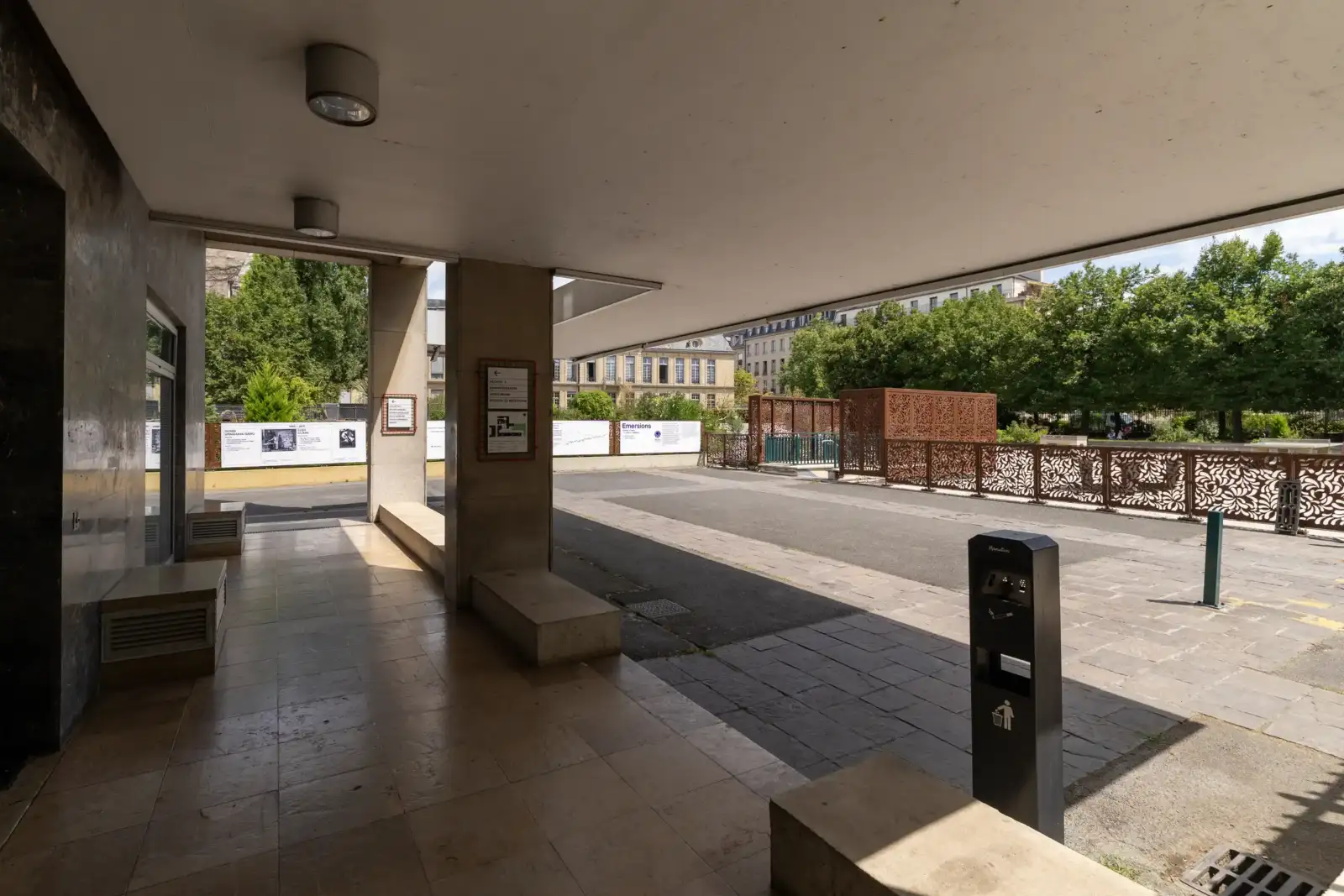 Vue ombragée depuis un porche moderne vers une cour pavée et des arbres.