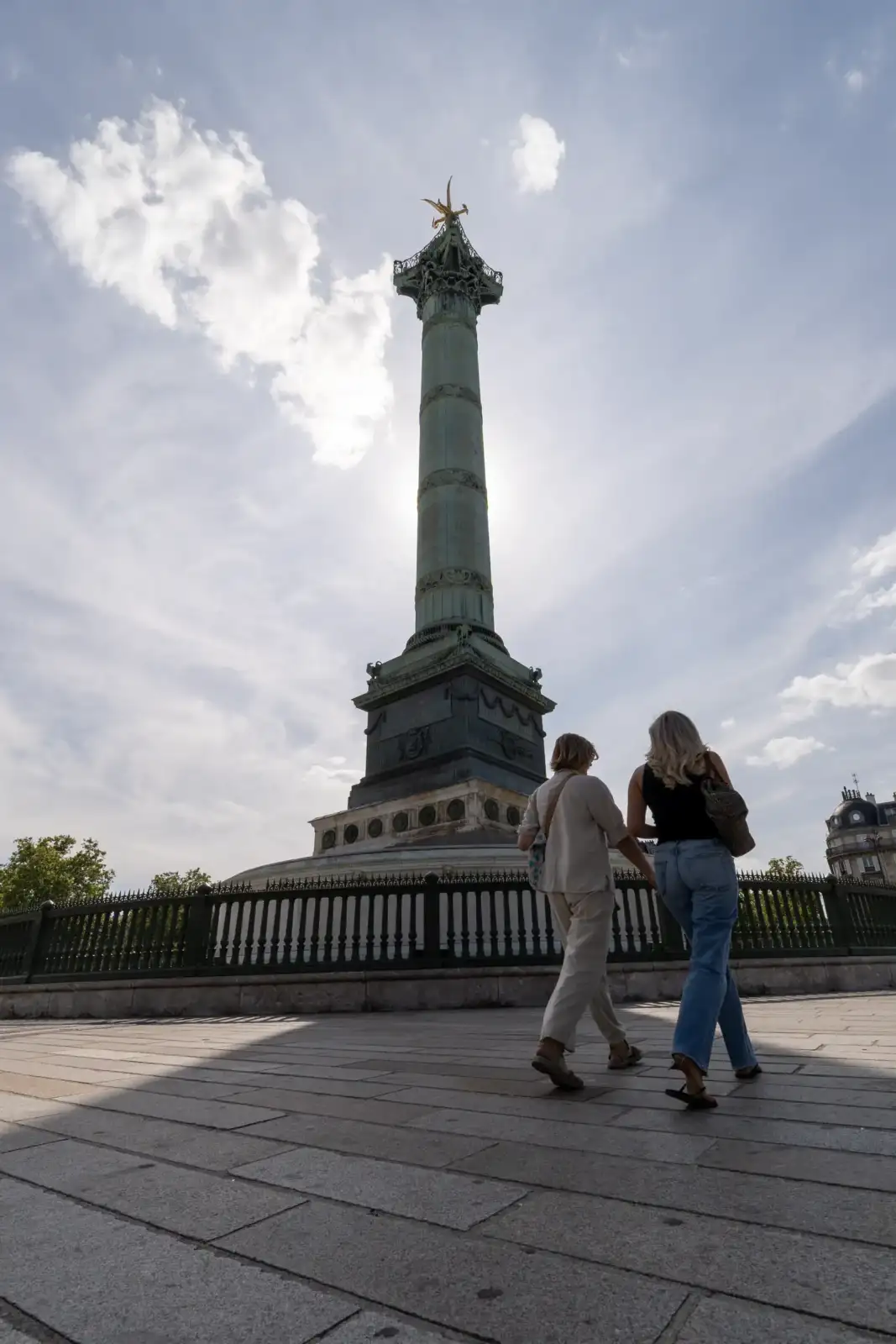 Deux femmes marchent devant la colonne de Juillet sur fond de ciel nuageux.