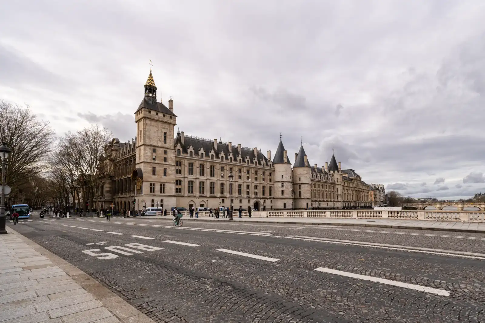 La façade historique de la Conciergerie borde une rue pavée sous un ciel nuageux.