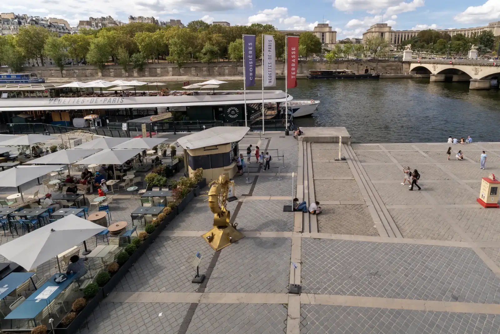 Vue plongeante des quais de Seine à Paris, montrant un bateau de croisière et une terrasse animée.