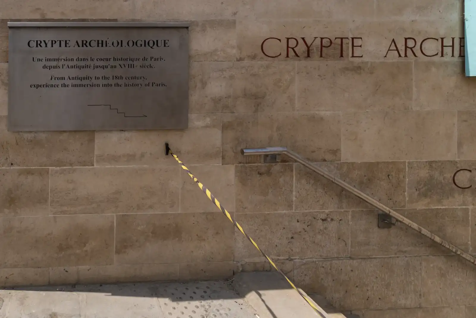 Mur en pierres claires affichant l'inscription de la crypte archéologique avec un panneau explicatif.