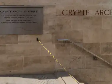 Mur en pierres claires affichant l'inscription de la crypte archéologique avec un panneau explicatif.