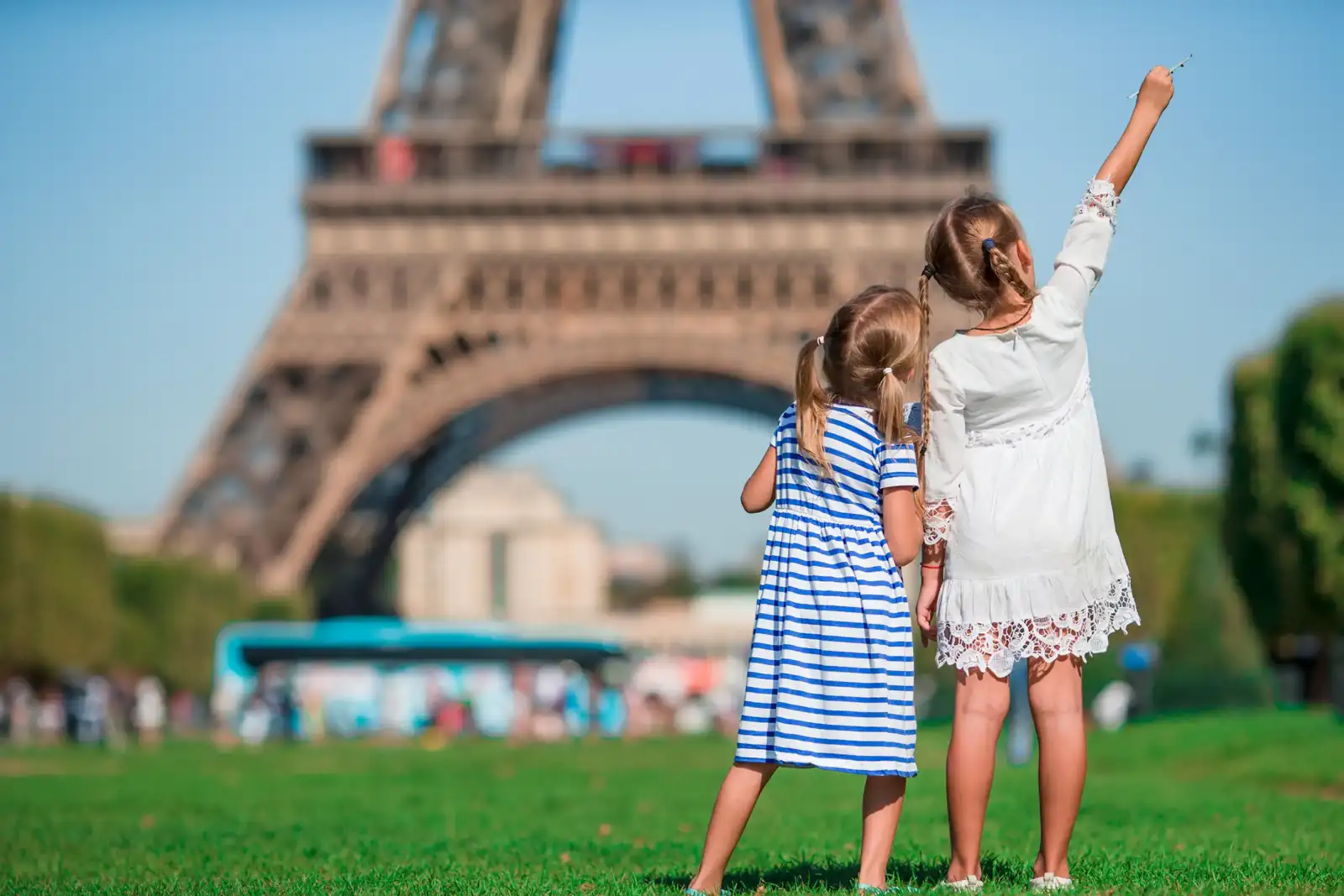 Deux petites filles regardent la Tour Eiffel dans un parc verdoyant sous un ciel bleu.