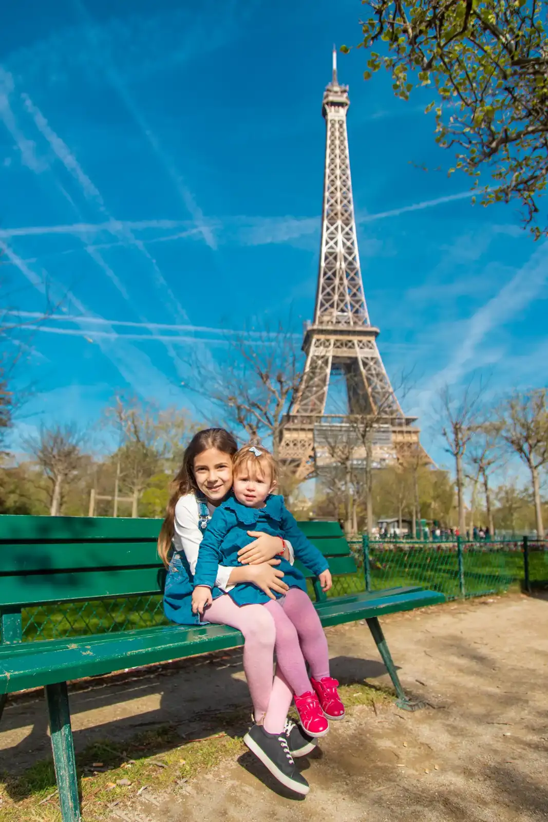 Deux fillettes sourient assises sur un banc vert avec la tour Eiffel en arrière-plan.