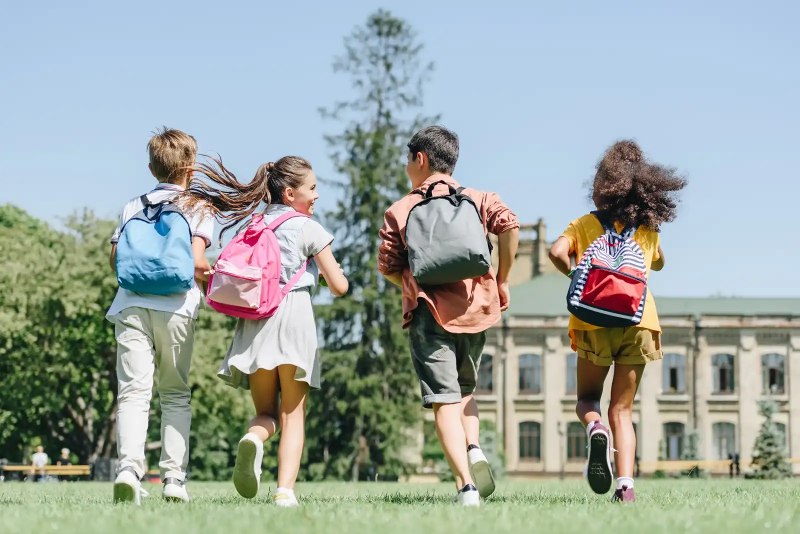Quatre écoliers avec des sacs à dos courent joyeusement dans un parc ensoleillé.