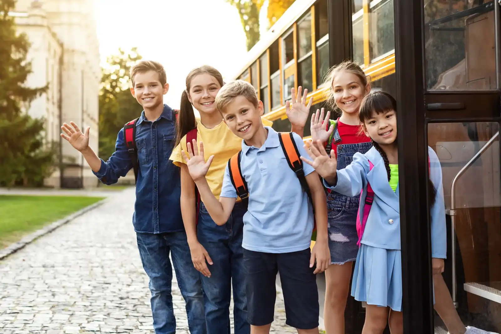 Des écoliers souriants font signe de la main devant un bus scolaire stationné à l extérieur.