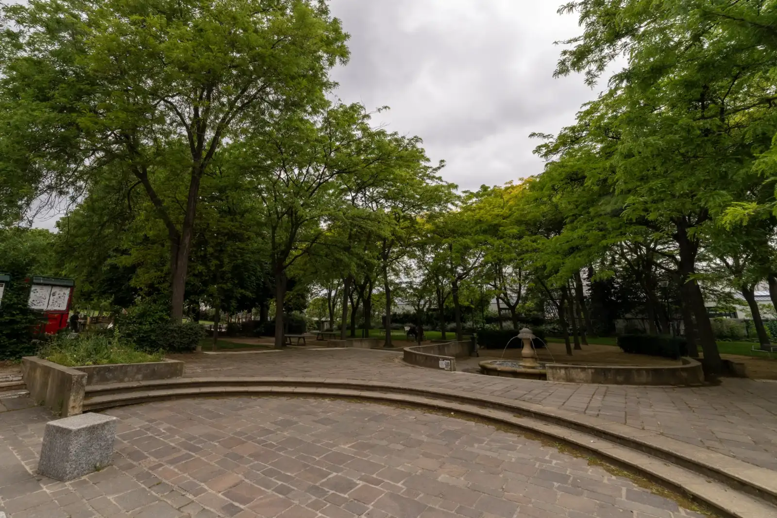 Une place pavée entourée d’arbres verdoyants sous un ciel couvert de nuages gris.