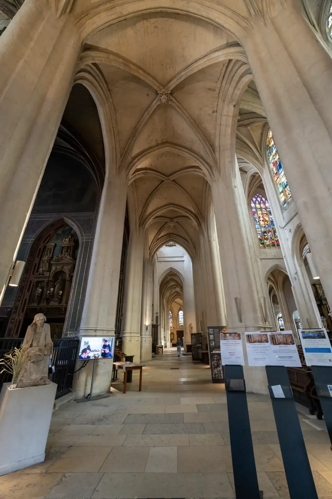 Vue intérieure de l'église Saint Gervais Saint Protais présentant des piliers gothiques et des panneaux explicatifs.