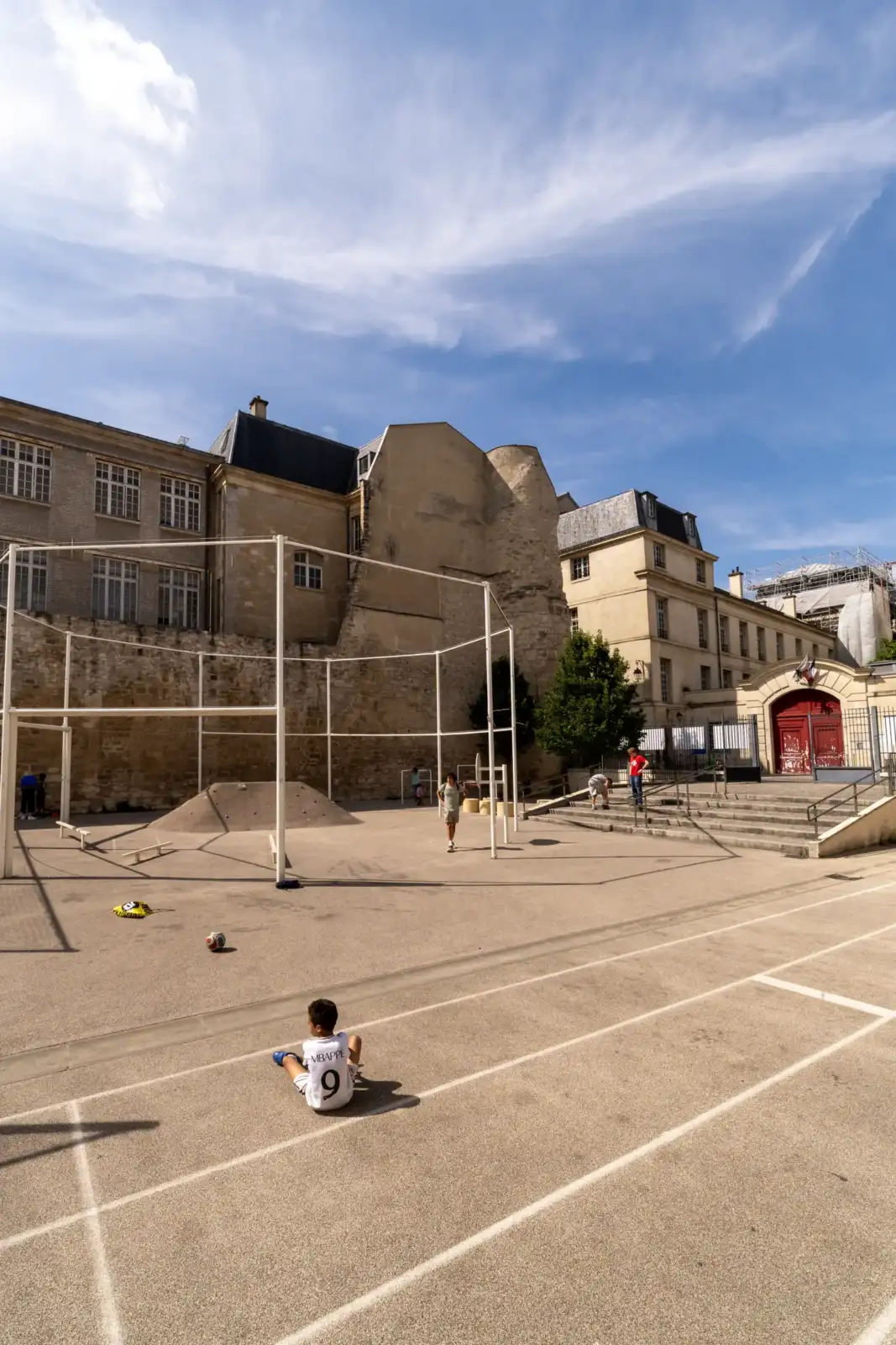 Un jeune garçon assis sur une cour d'école jouant au pied des vestiges de l'enceinte Philippe Auguste.