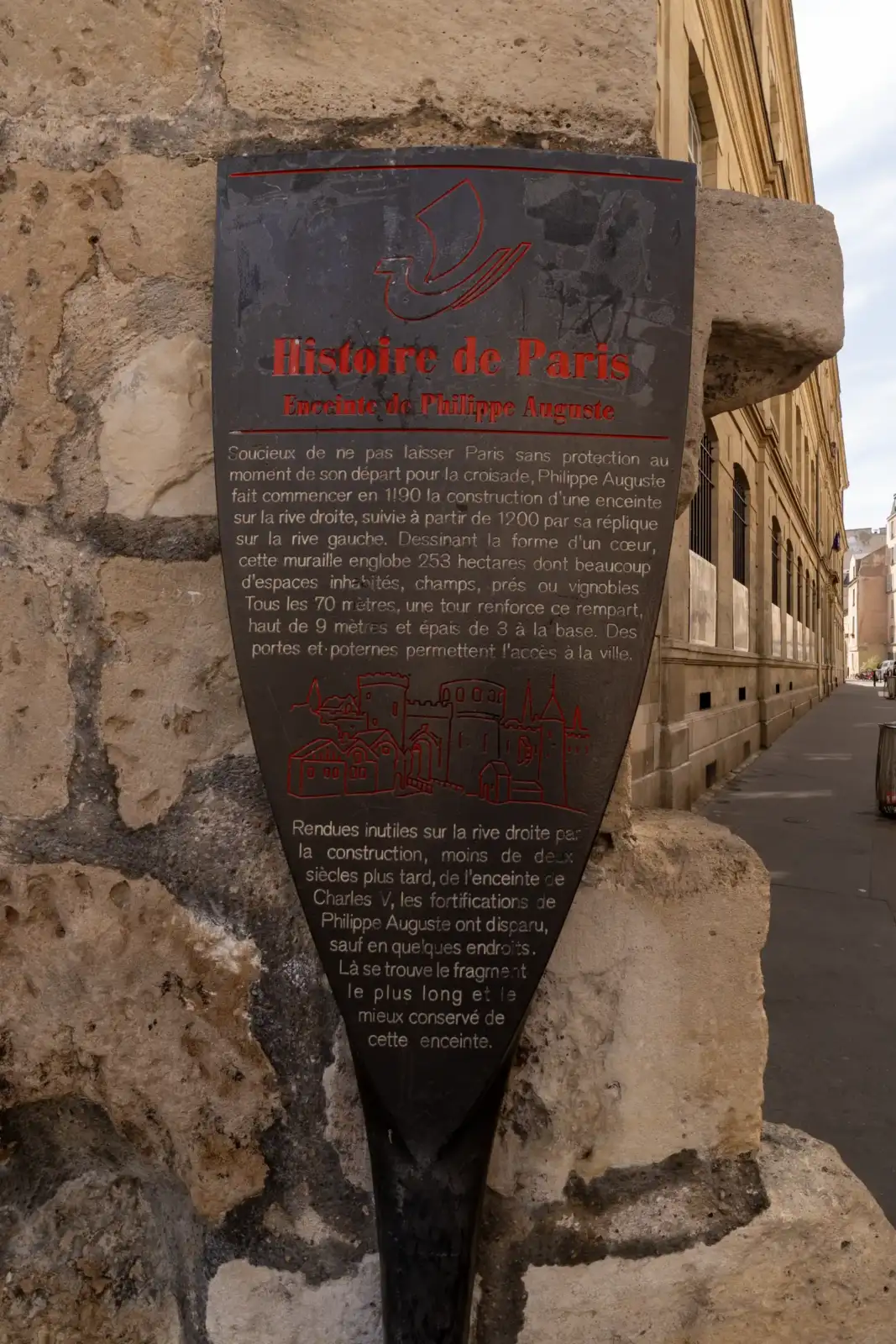 Panneau explicatif noir en forme de bouclier sur un vieux mur de pierre à Paris.