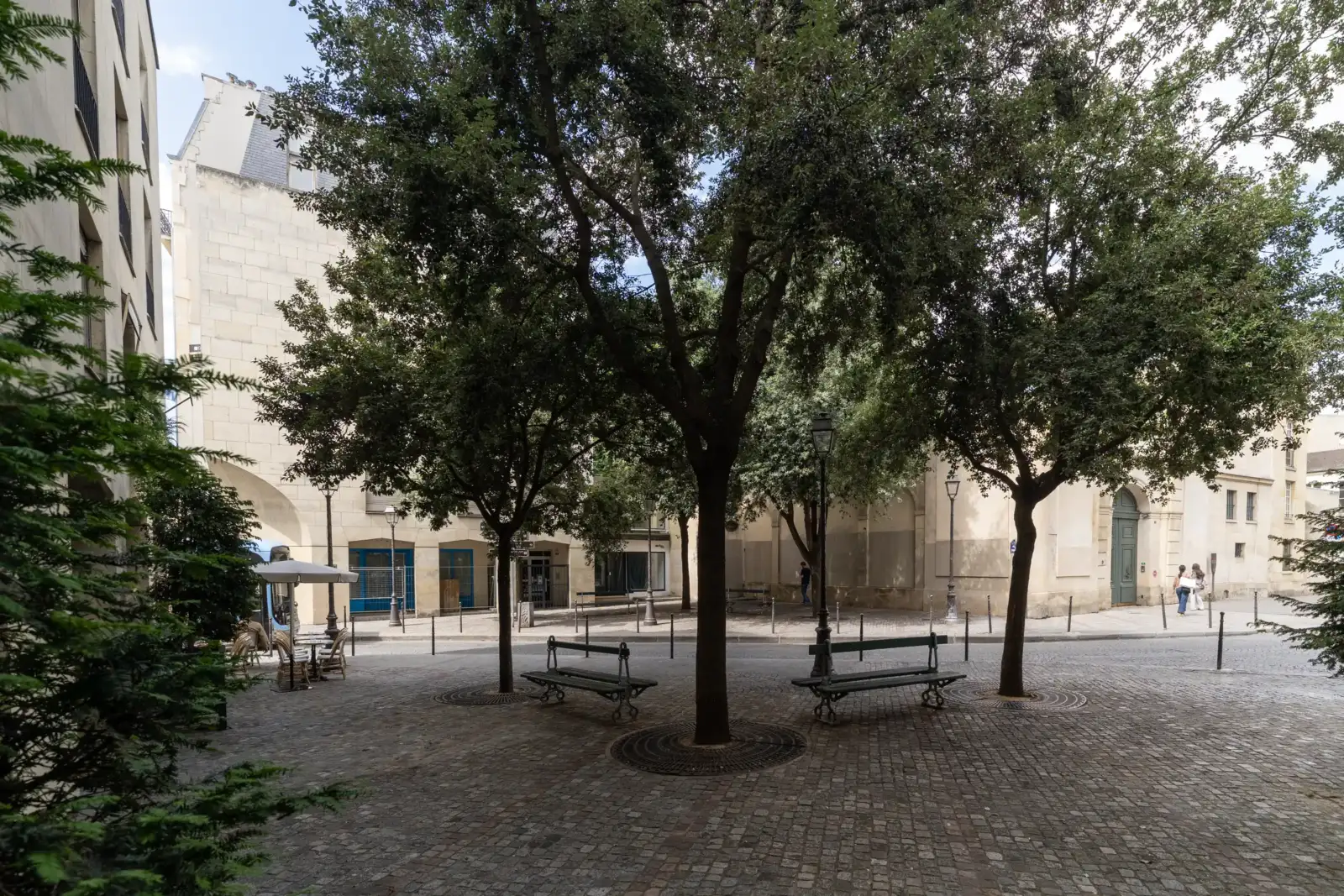 Une petite place parisienne pavée et ombragée entourée de bâtiments anciens et d'arbres.