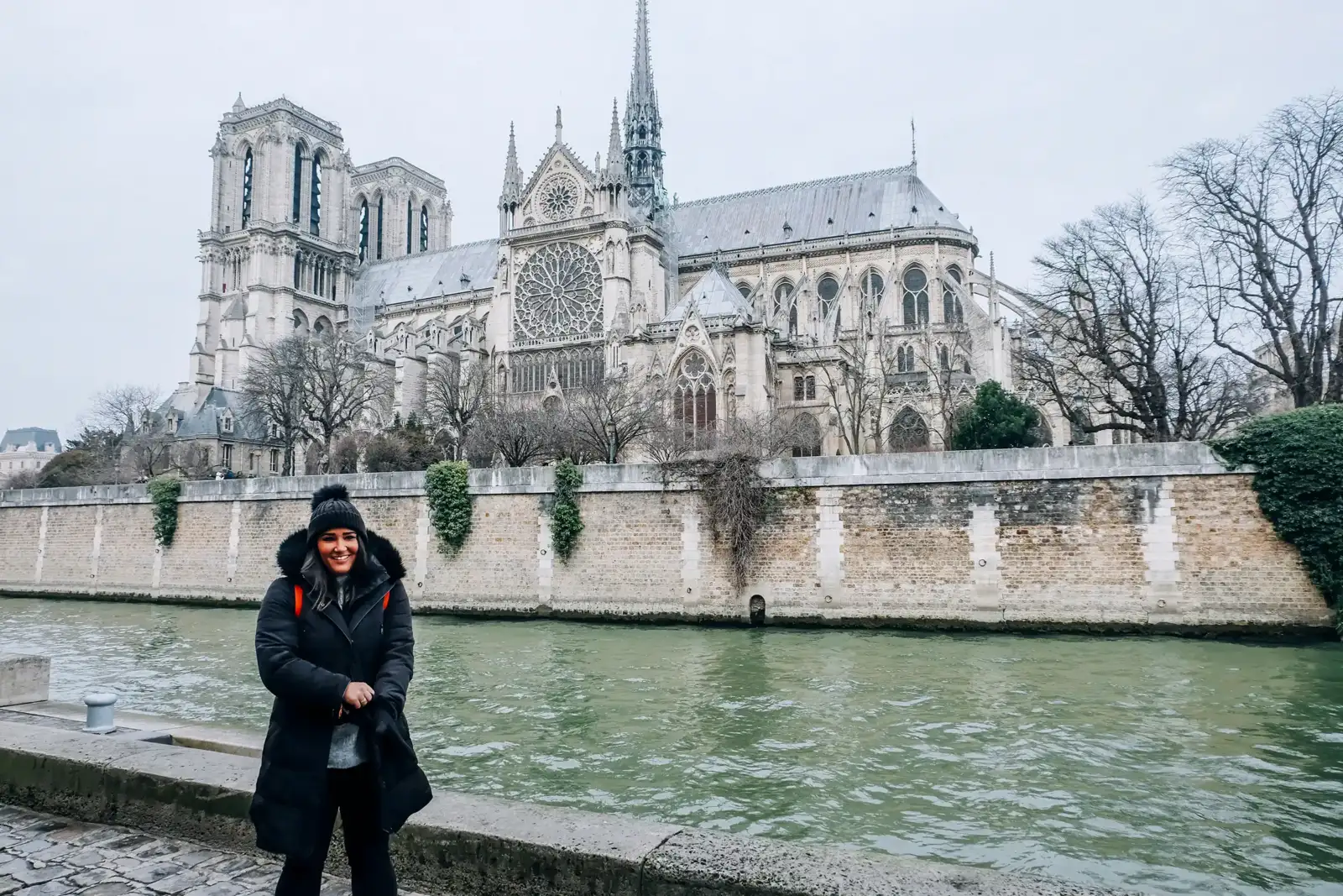 Une femme souriante en bonnet et manteau noir pose au bord de la Seine.