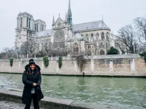 Une femme souriante en bonnet et manteau noir pose au bord de la Seine.