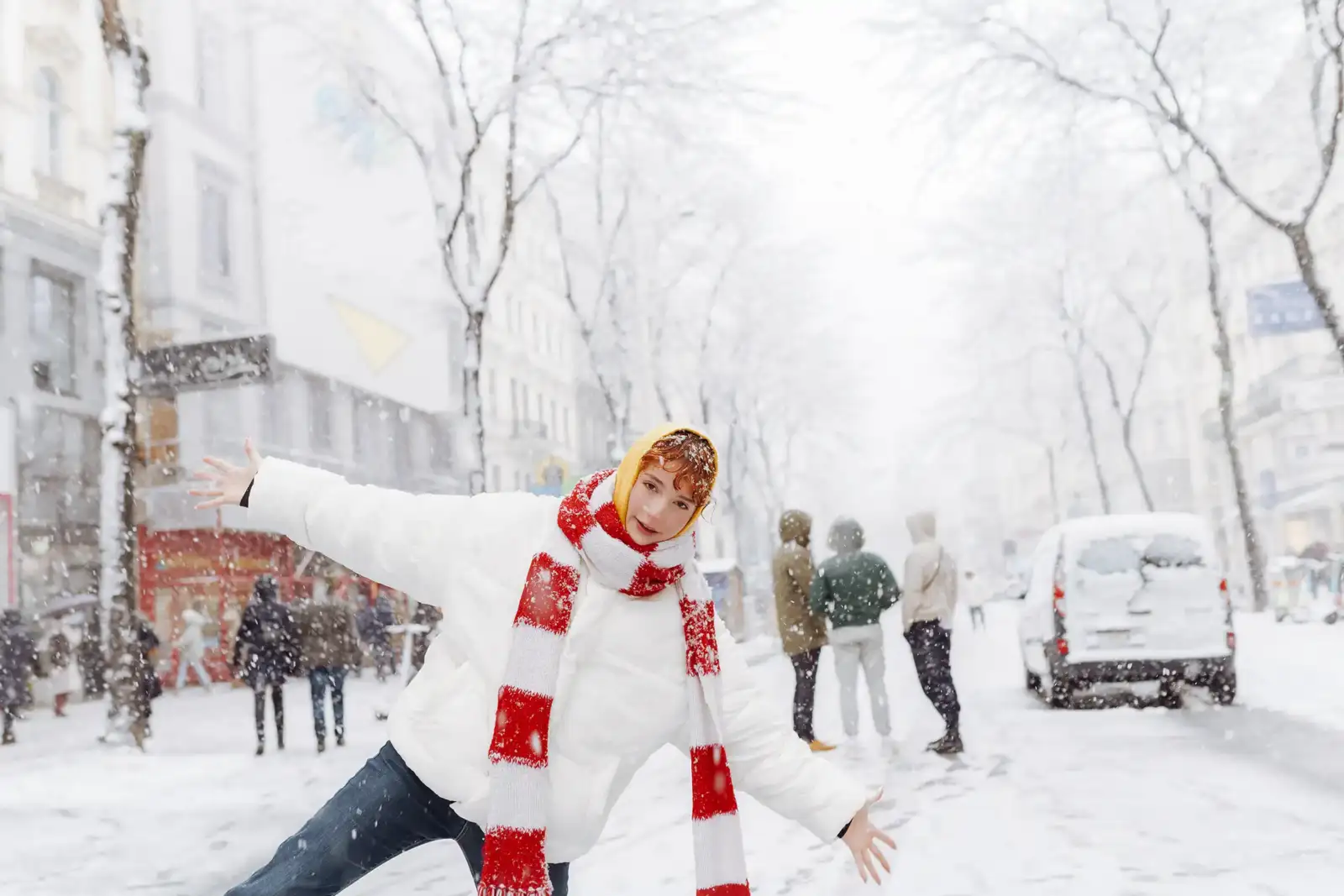 Une femme rousse en manteau blanc et écharpe rouge pose joyeusement sous la neige.