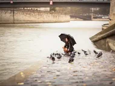 Une femme souriante sous un parapluie noir nourrit des pigeons au bord de la Seine.