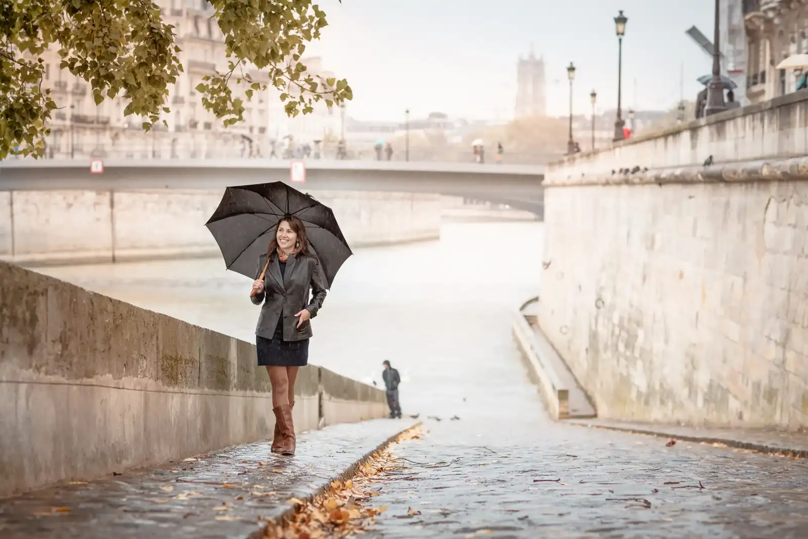Une femme sourit en marchant avec un parapluie noir sur les quais de Paris.