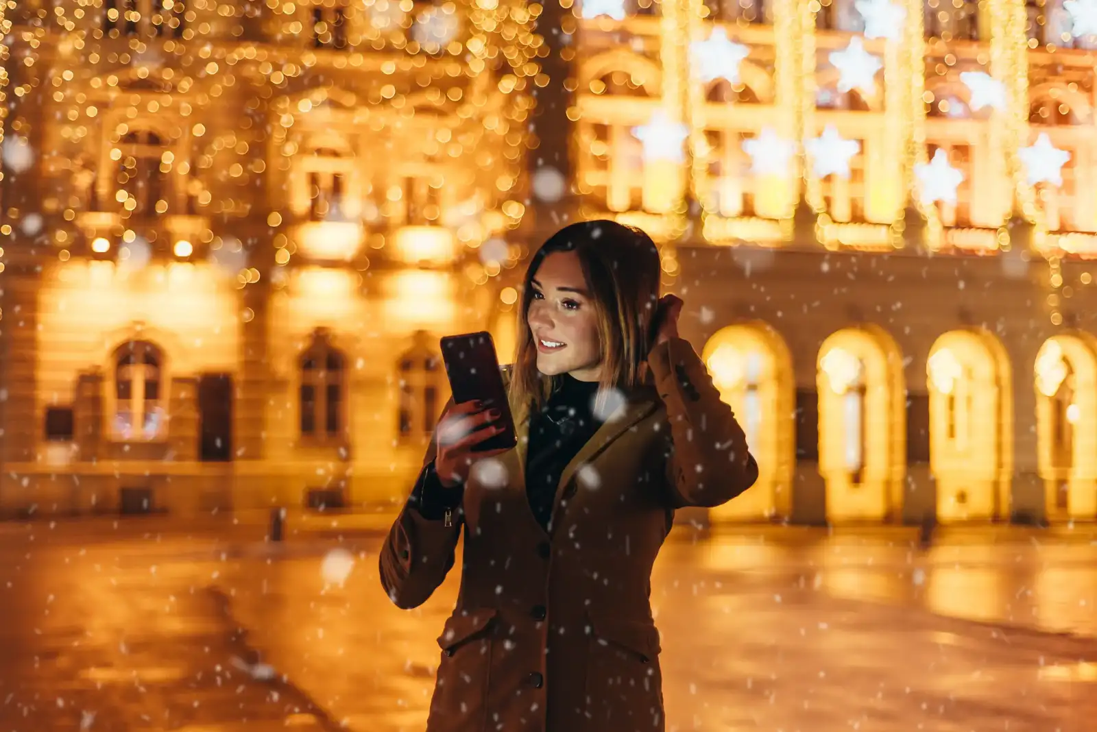 Une femme souriante regarde son smartphone devant un bâtiment parisien illuminé sous la neige.