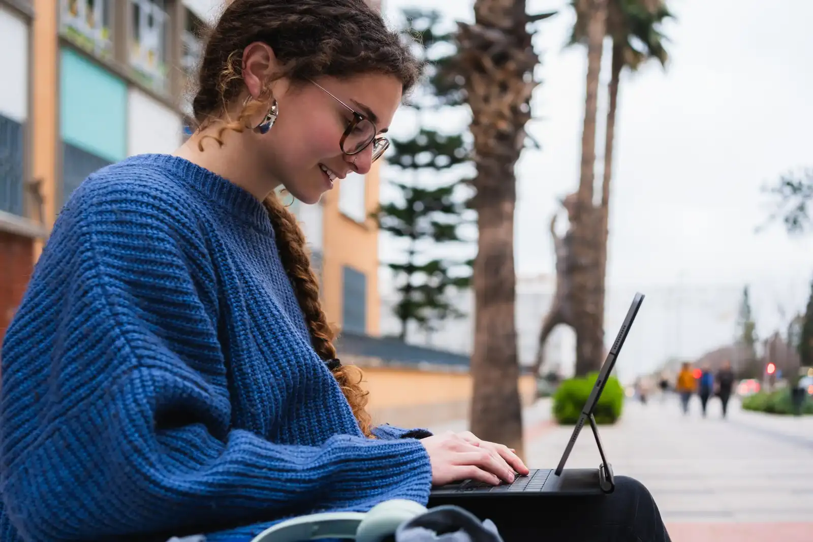Une jeune femme rousse avec des lunettes travaille sur une tablette numérique dehors.