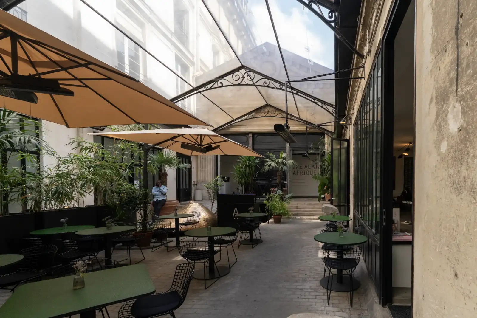 Une cour pavée et verrière abrite des tables vertes et de grands parasols beiges.