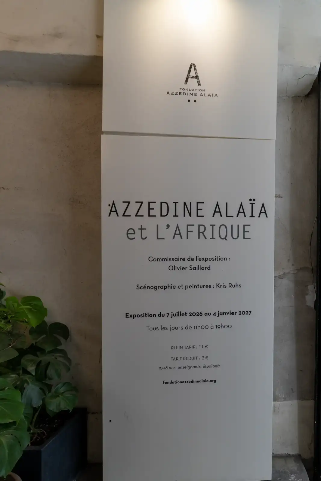 Un panneau blanc annonce l'exposition Azzedine Alaïa et l'Afrique devant un mur brut.