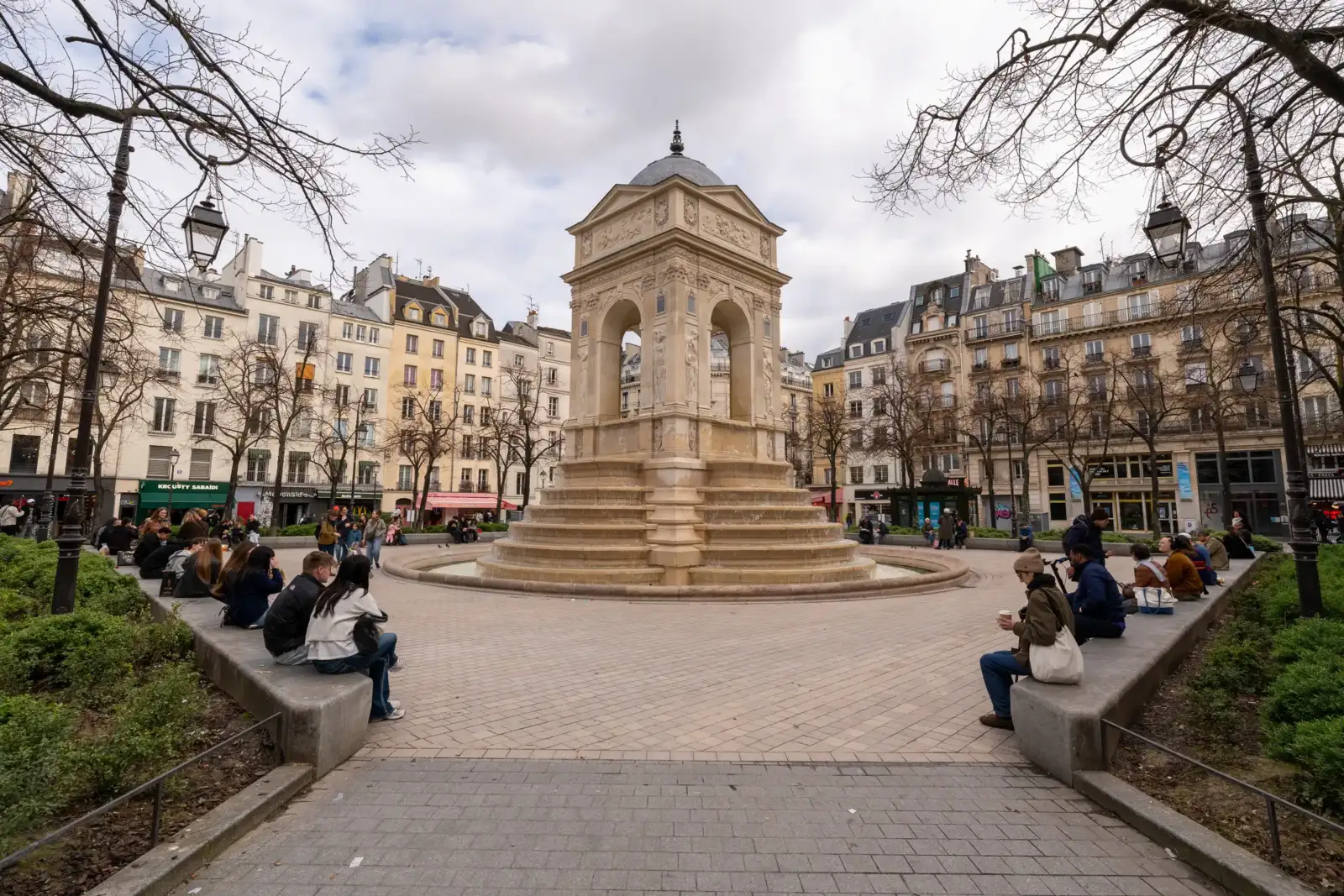 La fontaine des Innocents au centre d'une place parisienne entourée de nombreux passants assis.