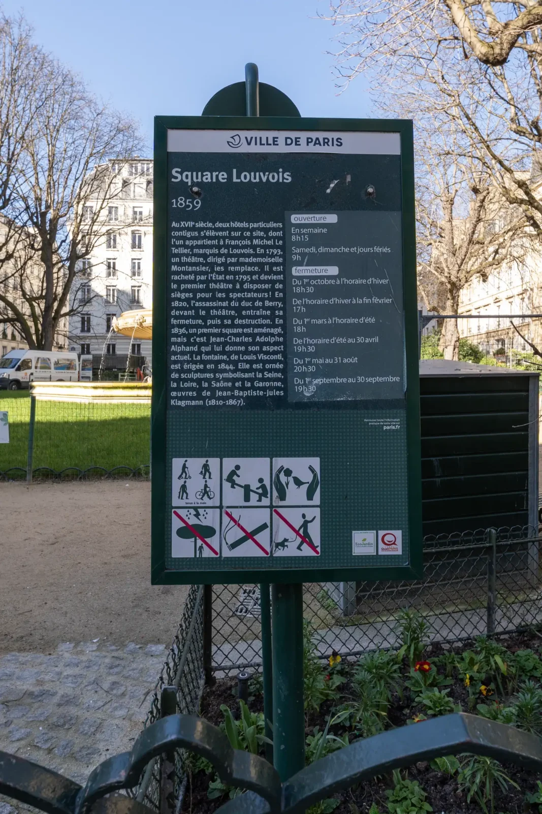 Panneau d'information vert sur le Square Louvois devant une fontaine dans un parc parisien.