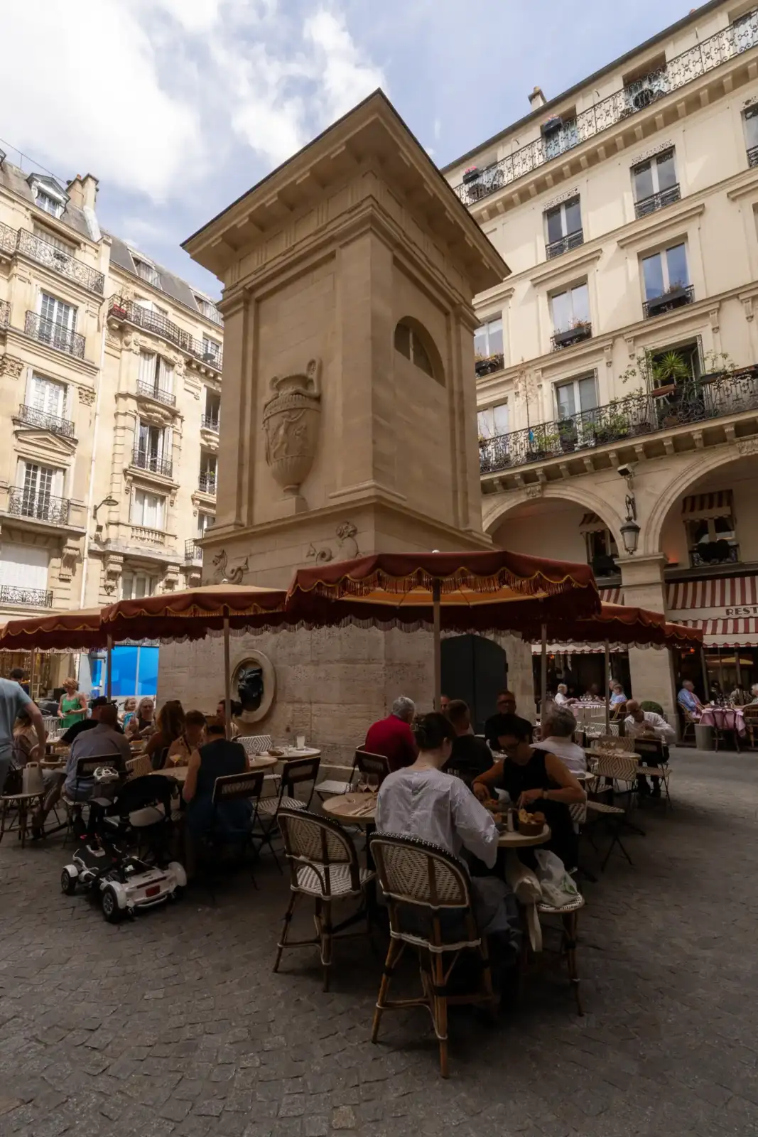 Une fontaine monumentale en pierre se dresse sur une place parisienne animée avec des terrasses.