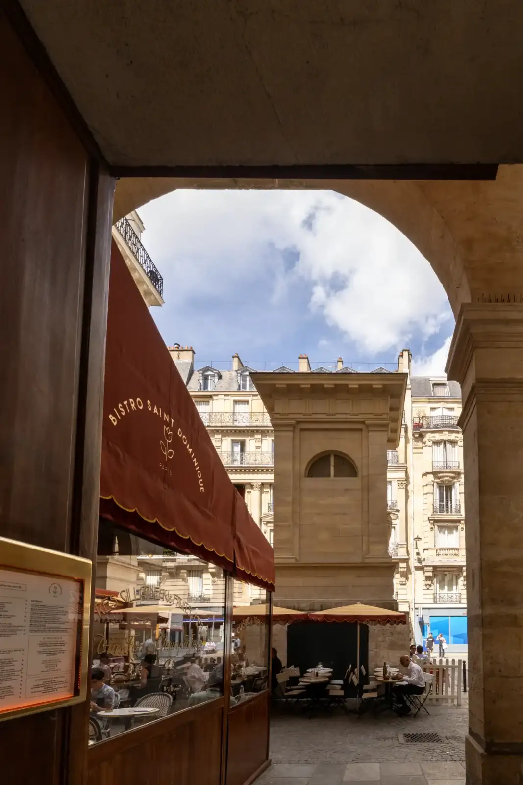 Vue encadrée d'une place parisienne avec un bistro et la fontaine de Mars sous un ciel nuageux.