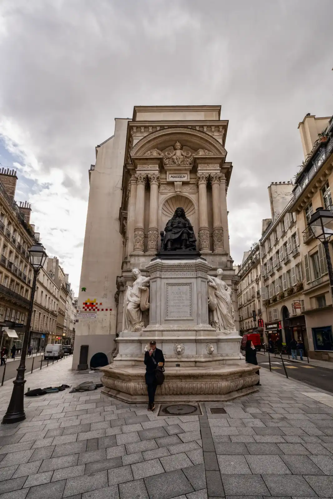 La monumentale fontaine Molière se dresse entre deux immeubles parisiens sous un ciel nuageux.
