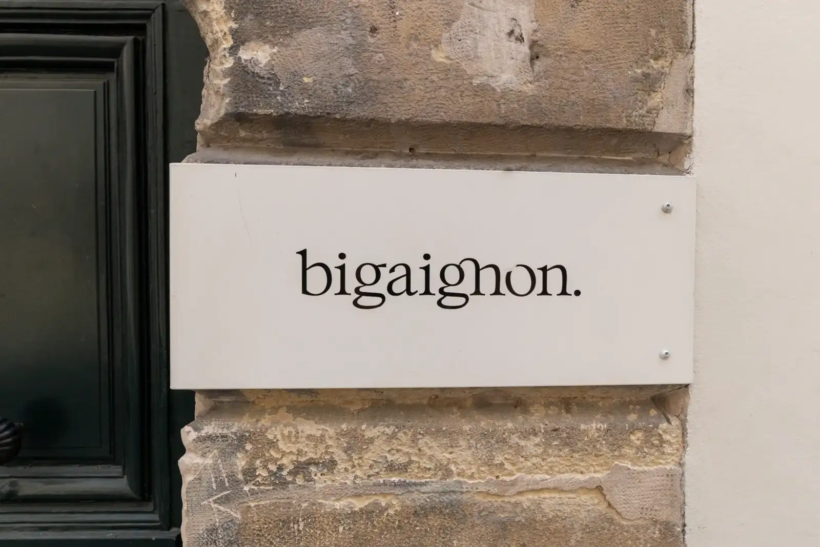 Un panneau rectangulaire blanc affiche le nom bigaignon sur un mur en pierre.