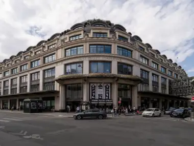 Façade élégante du grand magasin parisien sous un ciel nuageux avec quelques voitures.