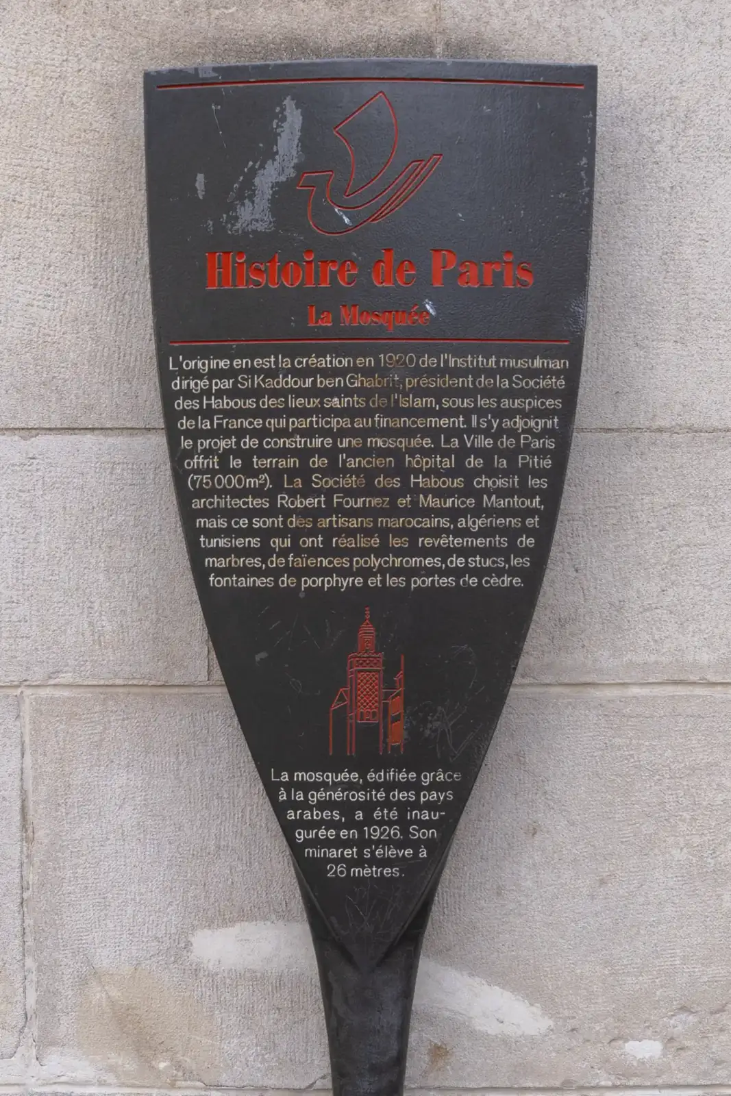 Panneau noir d’information historique en métal fixé sur un mur en pierre grise.