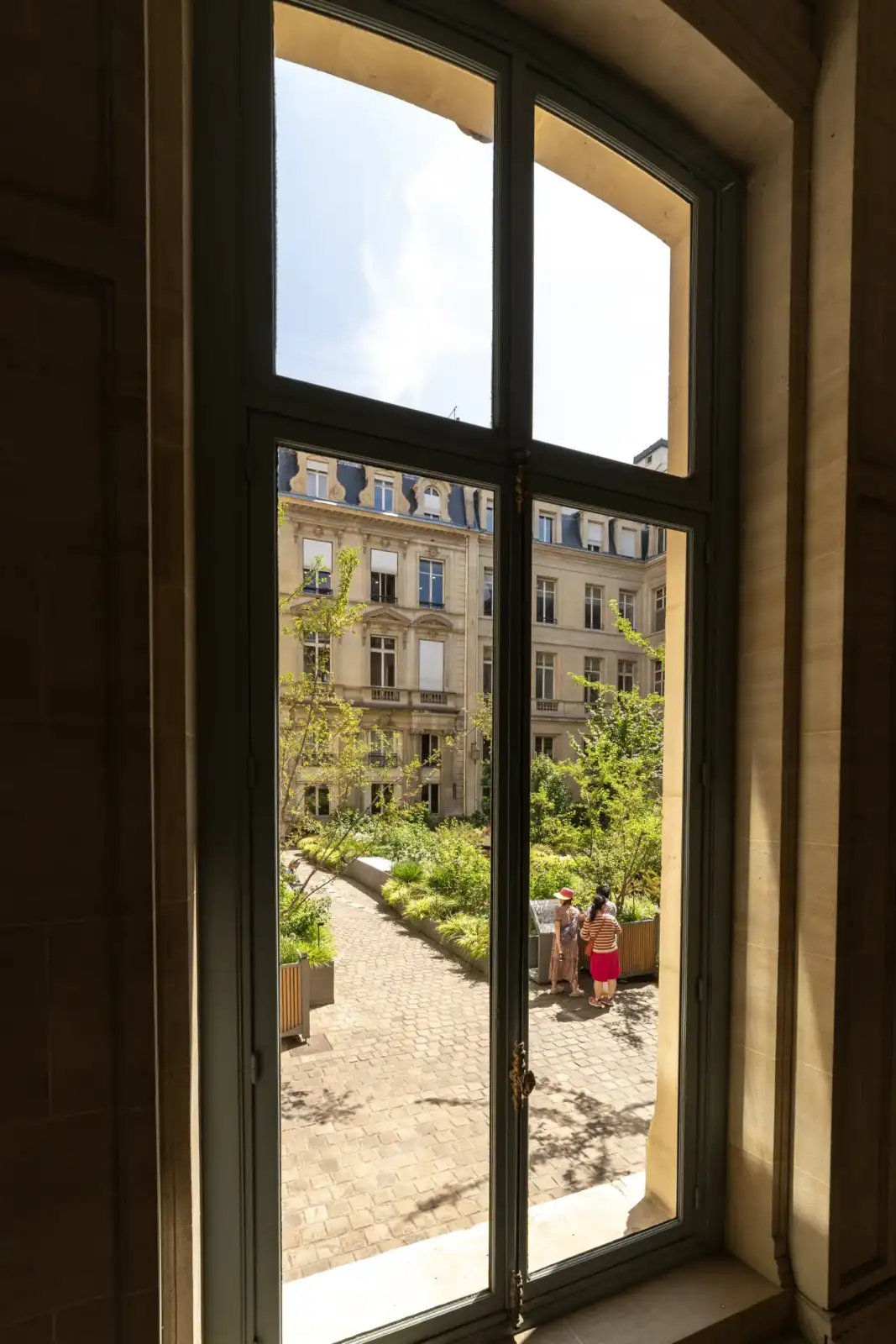 Une grande fenêtre en bois gris offre une vue sur une cour pavée ensoleillée.